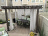A wooden pergola with white curtains enhances the backyard, anchored by galvanized Titan post anchors. It features a dark rectangular table with six green cushioned chairs. Yellow flowers and a light wood deck adorn the space, set against beige houses, all complimented by a 4x4 Triple Bundle - White/Black.