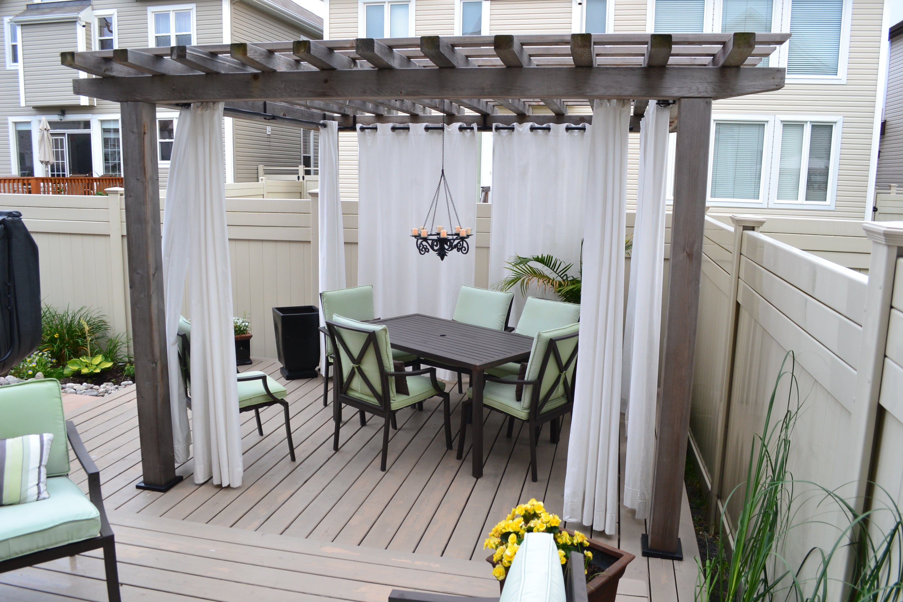 A wooden pergola with sheer white curtains graces the patio, secured by the Post Anchor Single. Beneath, a dark dining table with six green-cushioned chairs sits under a black chandelier. The area is enclosed by beige fencing, and yellow flowers add brightness to the scene.