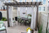 A wooden pergola with sheer white curtains graces the patio, secured by the Post Anchor Single. Beneath, a dark dining table with six green-cushioned chairs sits under a black chandelier. The area is enclosed by beige fencing, and yellow flowers add brightness to the scene.
