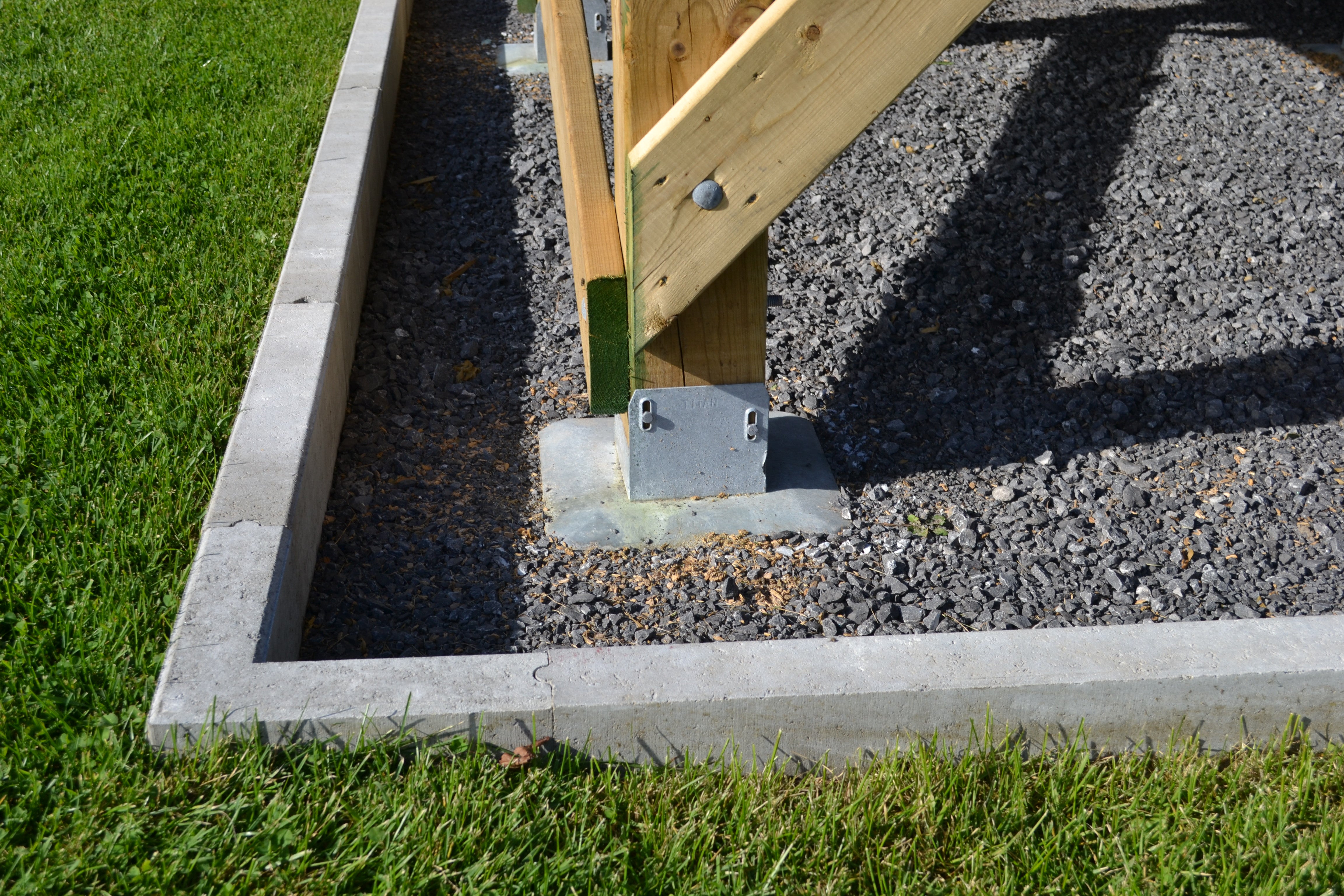 Close-up of a wooden post secured with a metal bracket and 1/4x3 Galv. Hex Head Lag Screws (4 pcs) on a gravel base, surrounded by a concrete border on grass.