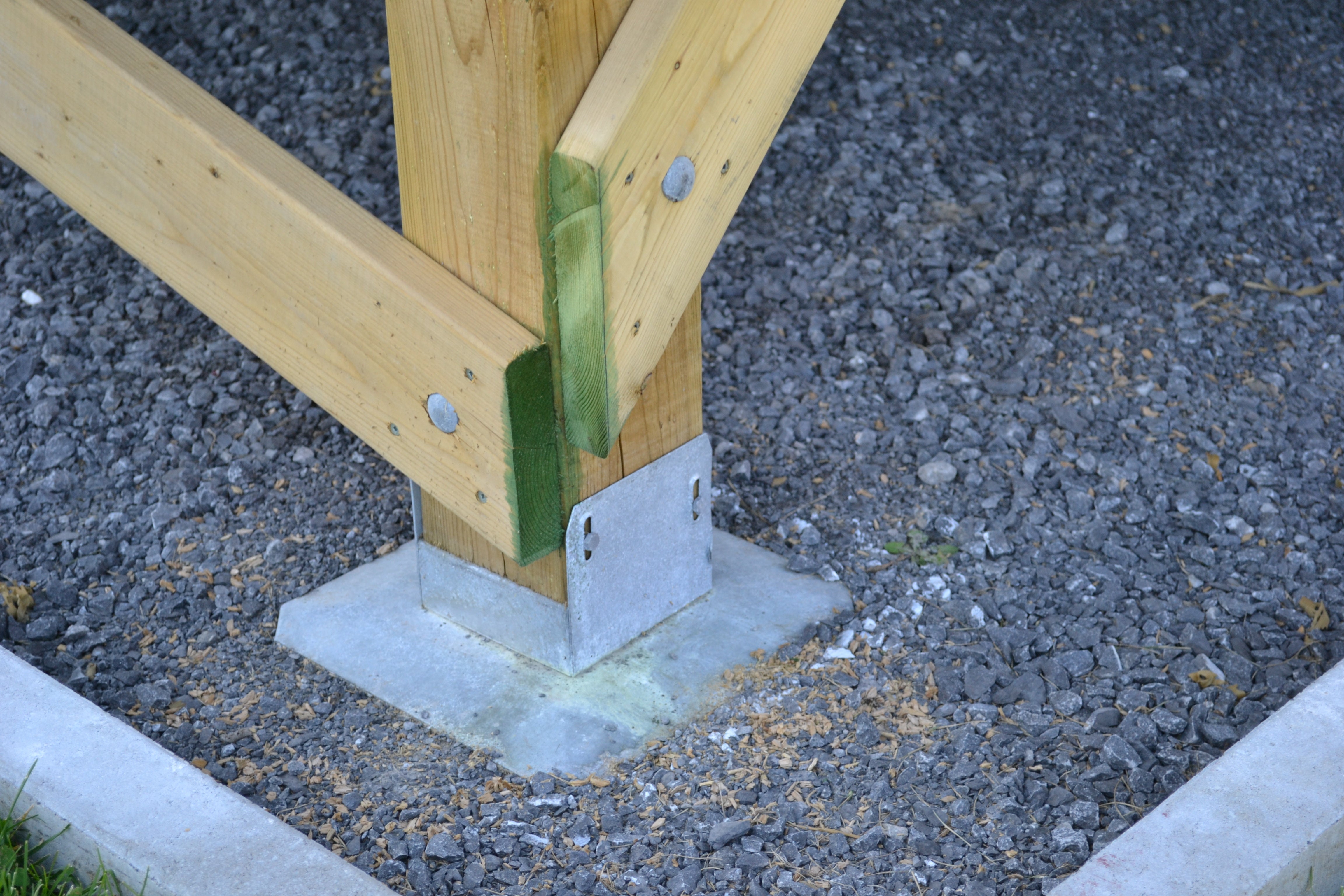 A wooden beam with a green-painted base is securely fastened to a metal bracket and cement base set on gravel using 1/4x3 Galv. Hex Head Lag Screws (4 pcs). Scattered wood shavings suggest recent activity at the site.