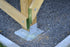 A wooden beam with a green-painted base is securely fastened to a metal bracket and cement base set on gravel using 1/4x3 Galv. Hex Head Lag Screws (4 pcs). Scattered wood shavings suggest recent activity at the site.
