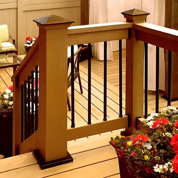 The wooden deck features black metal railings fastened with #14x4 Black Flat Head Screws. Red and white flowers in planters add charm, while a chair and table create a cozy atmosphere, all supported by sturdy Titan Wood Post Anchors.