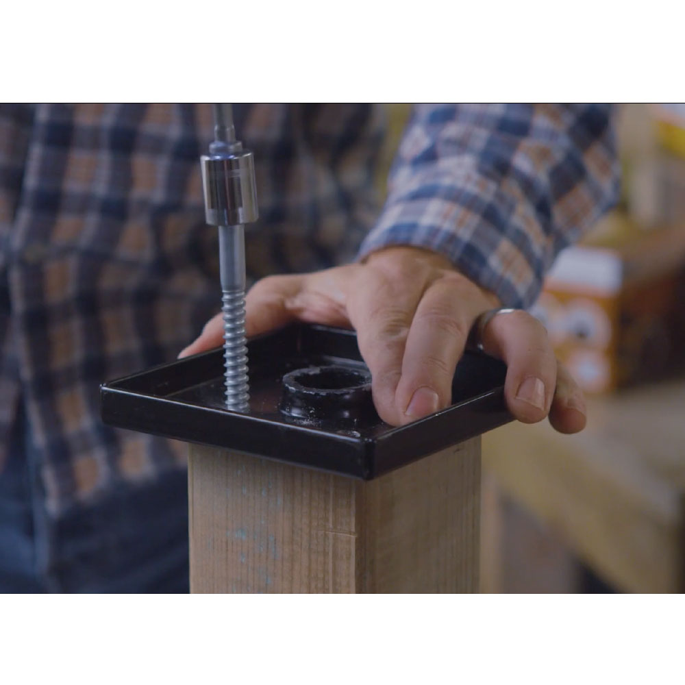 A person in a plaid shirt uses 3/8x5 Galv. Hex Head Lag Screws to attach a metal bracket to the top of a wooden post, holding the bracket with their left hand and operating the drill with their right hand, driving the screw into the Hot Dip Galvanized Titan Wood Post Anchor.