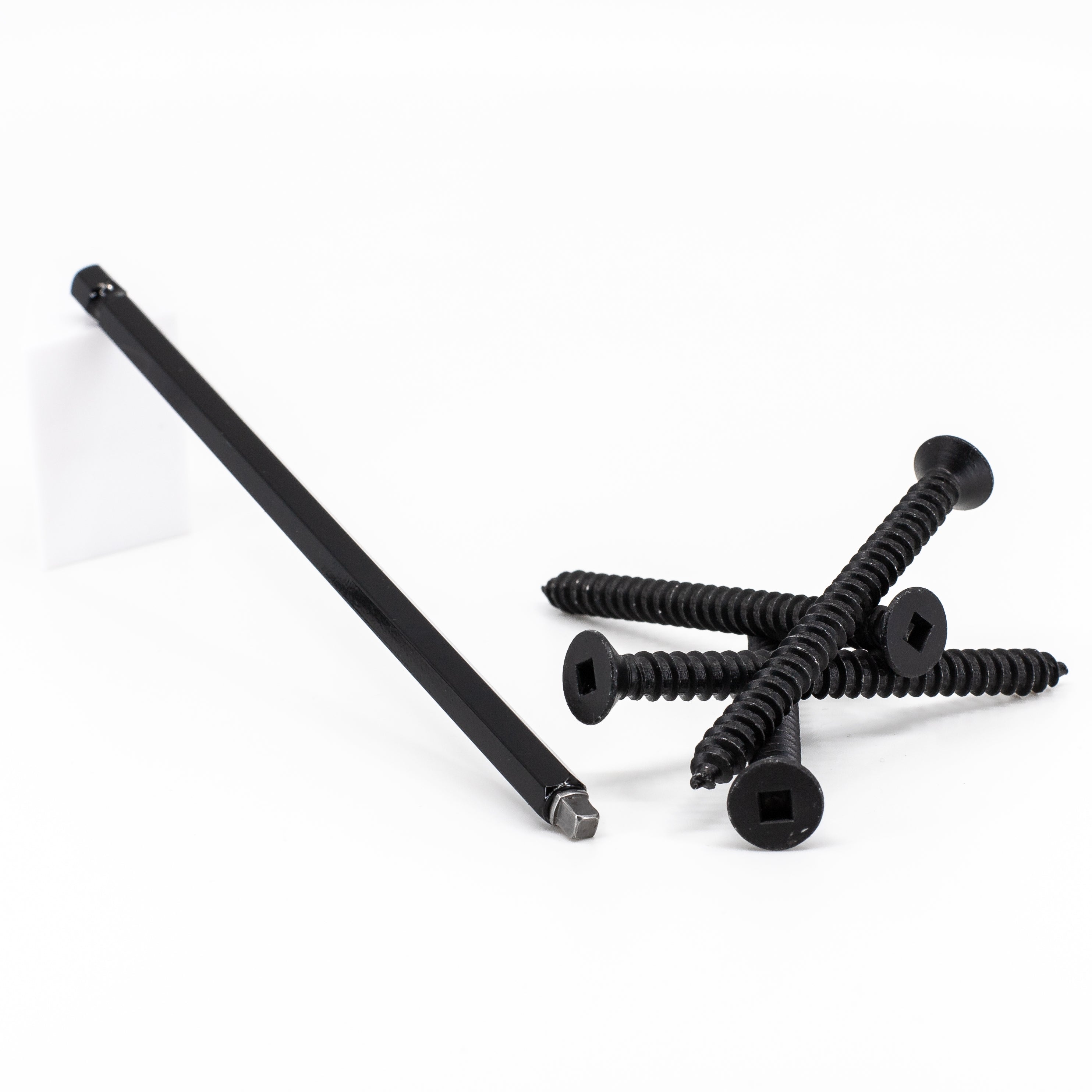 A black drill bit and four black square-head screws are casually arranged on a white surface. They ensure easy installation when used with the #3 Square Six Inch Driver Bit for precision and efficiency.