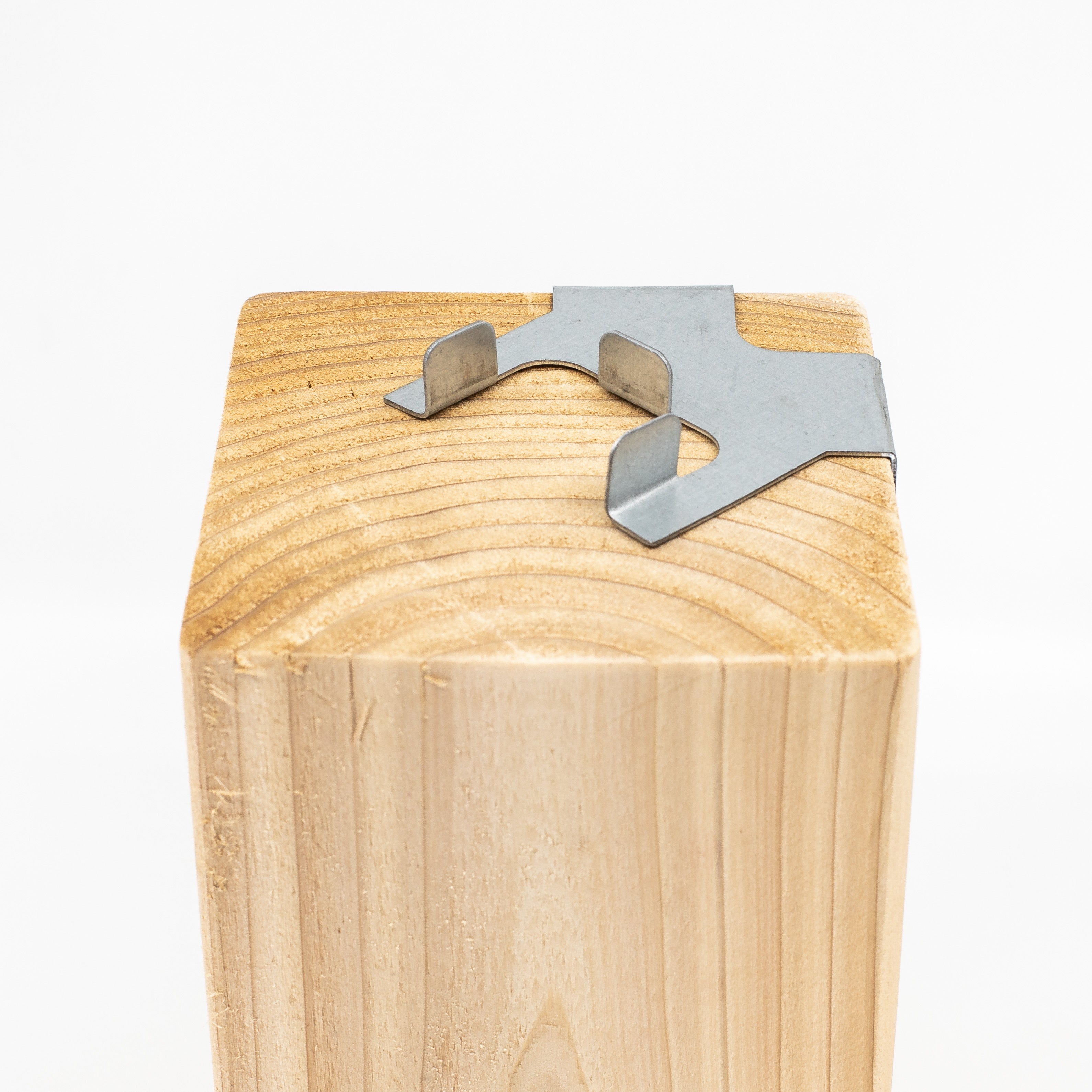 The Center Guide for Hole Saws displays a light wood grain texture with angled shapes. A metal bracket with protruding sections securely tops the wooden block, resembling a wood post anchor, all set against a plain white background that highlights its simplicity.