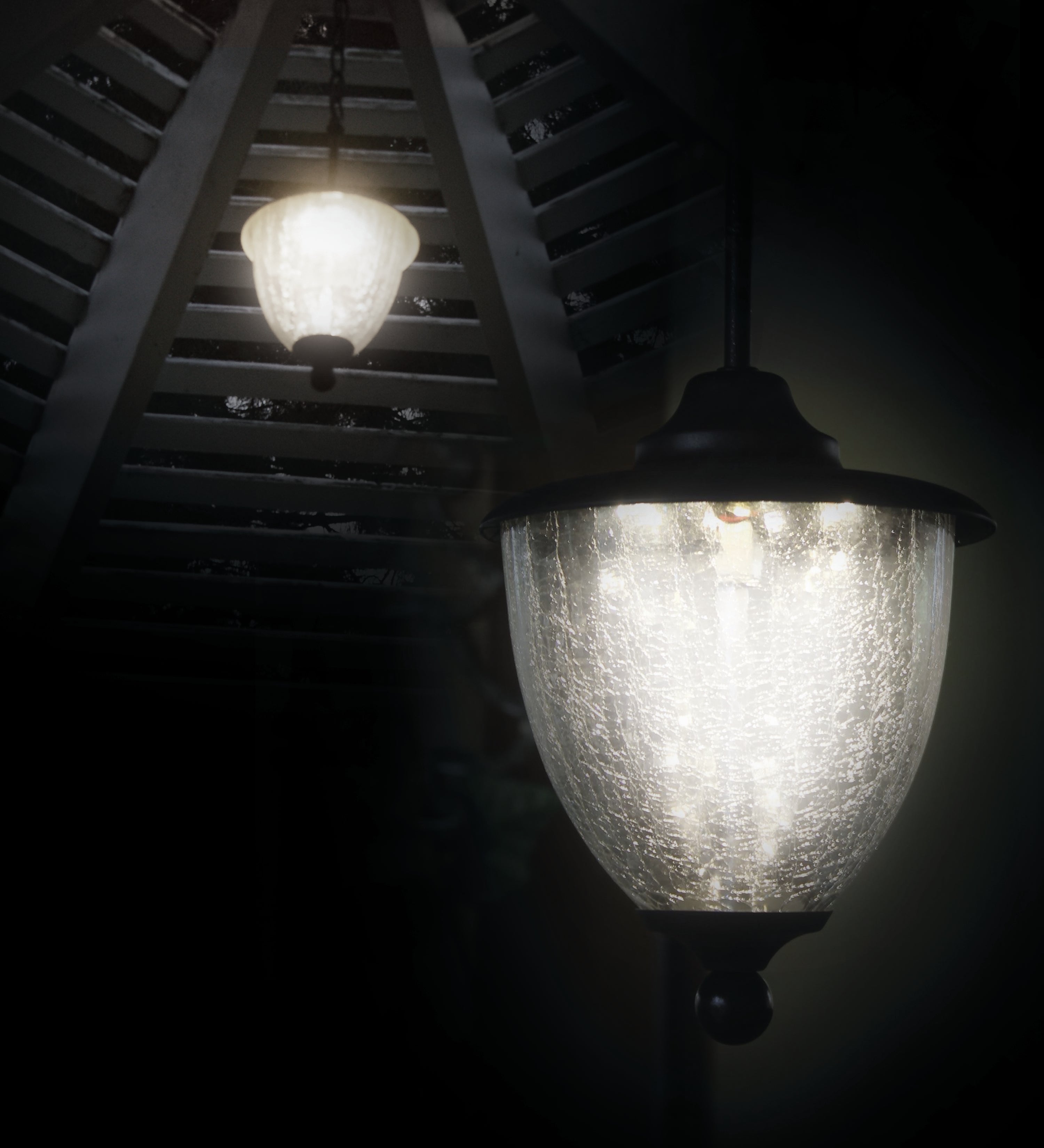 Two Classy Eclipse Solar Hanging Chandeliers adorn the decorative slatted wood ceiling, enhancing the outdoor decor. The nearest chandelier has an LED crackled glass cover, while the other is further away and dimmer, casting a soft glow in the dark.
