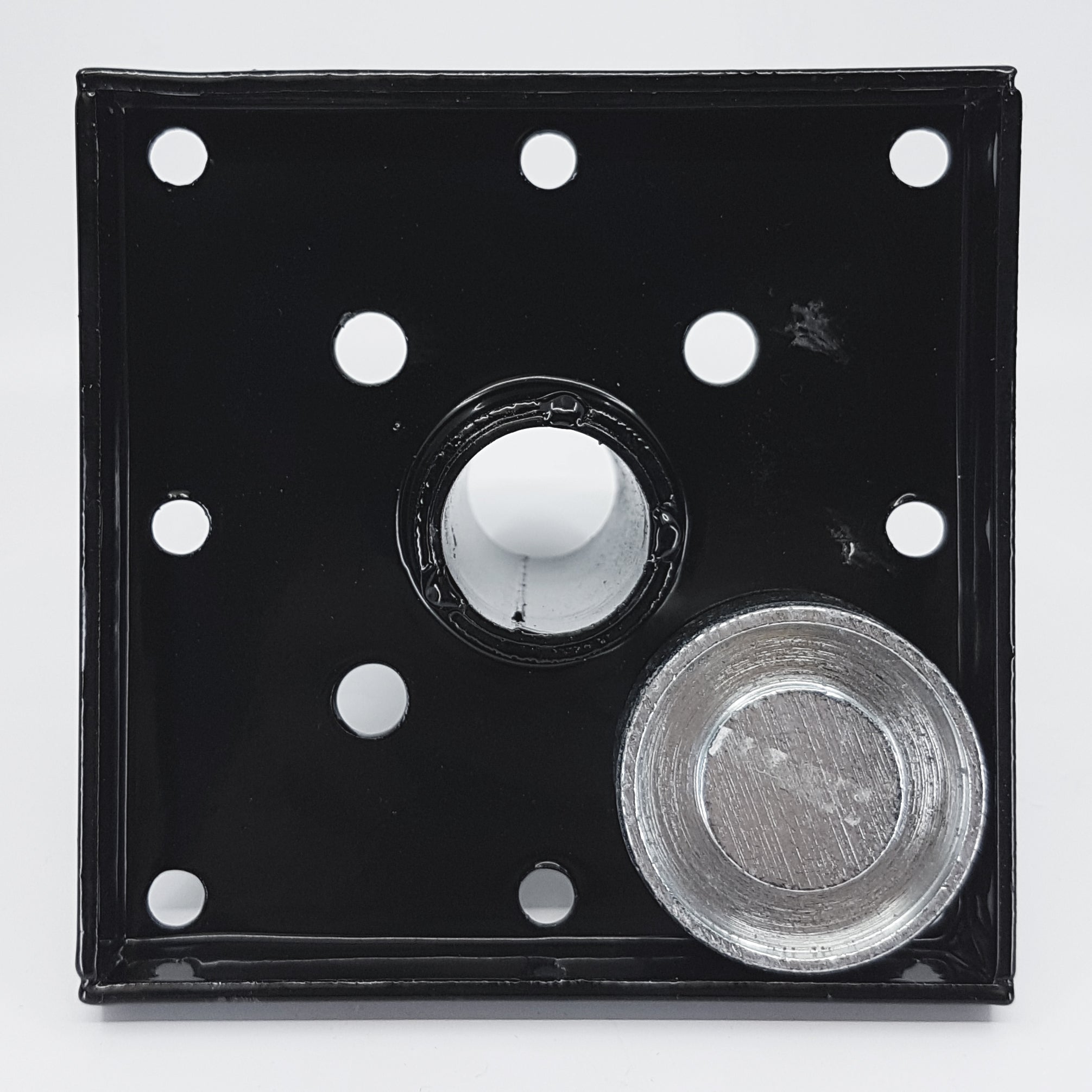The Hammer Tool - 44 is a black square metal plate with eight small holes and a central hole ideal for 4x4 post anchors. A recessed-center circular silver metal piece is next to it, designed for builders, all set against a plain white background.