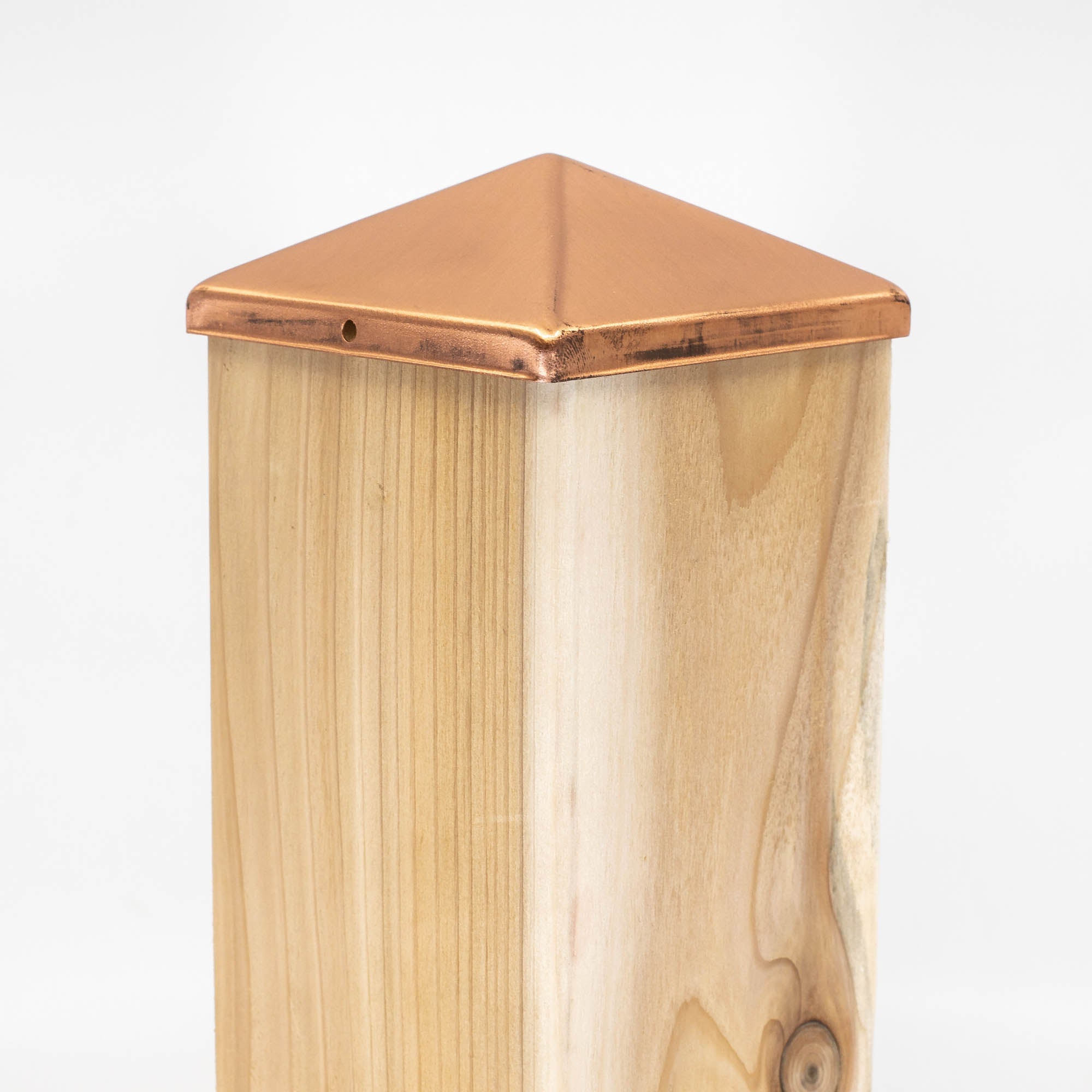 A wooden post adorned with a Pyramid Metal Post Cap made of copper highlights the wood grain through its natural finish. The cap features a small side hole, all presented on a plain white background for a sleek and modern appearance.