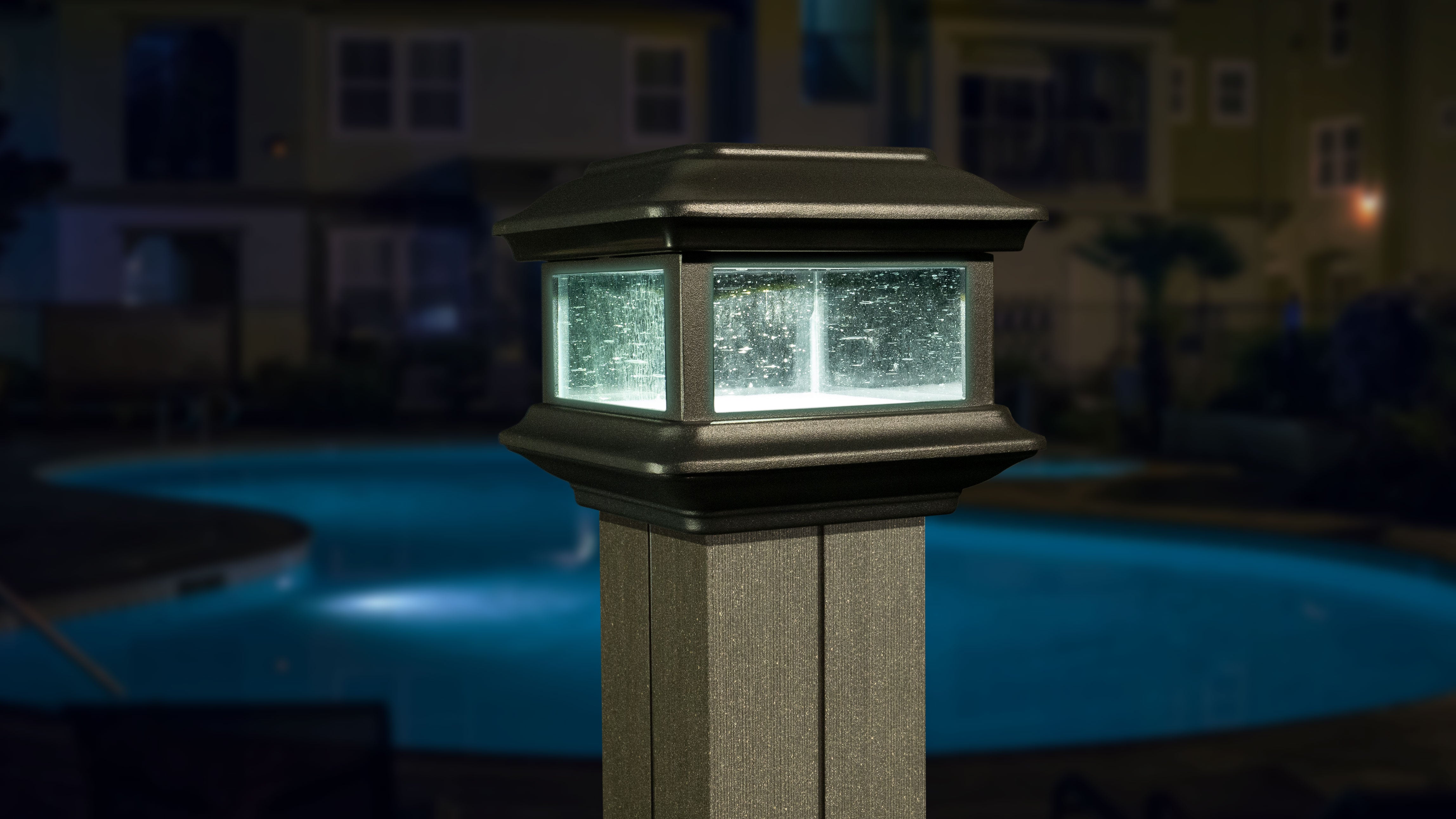A Classy Post Cap Colonial Solar lamp with pebbled glass panels stands on a post, illuminating the scene. In the background, a swimming pool reflects night lights while residential buildings peek through. The atmosphere is serene and dimly lit.