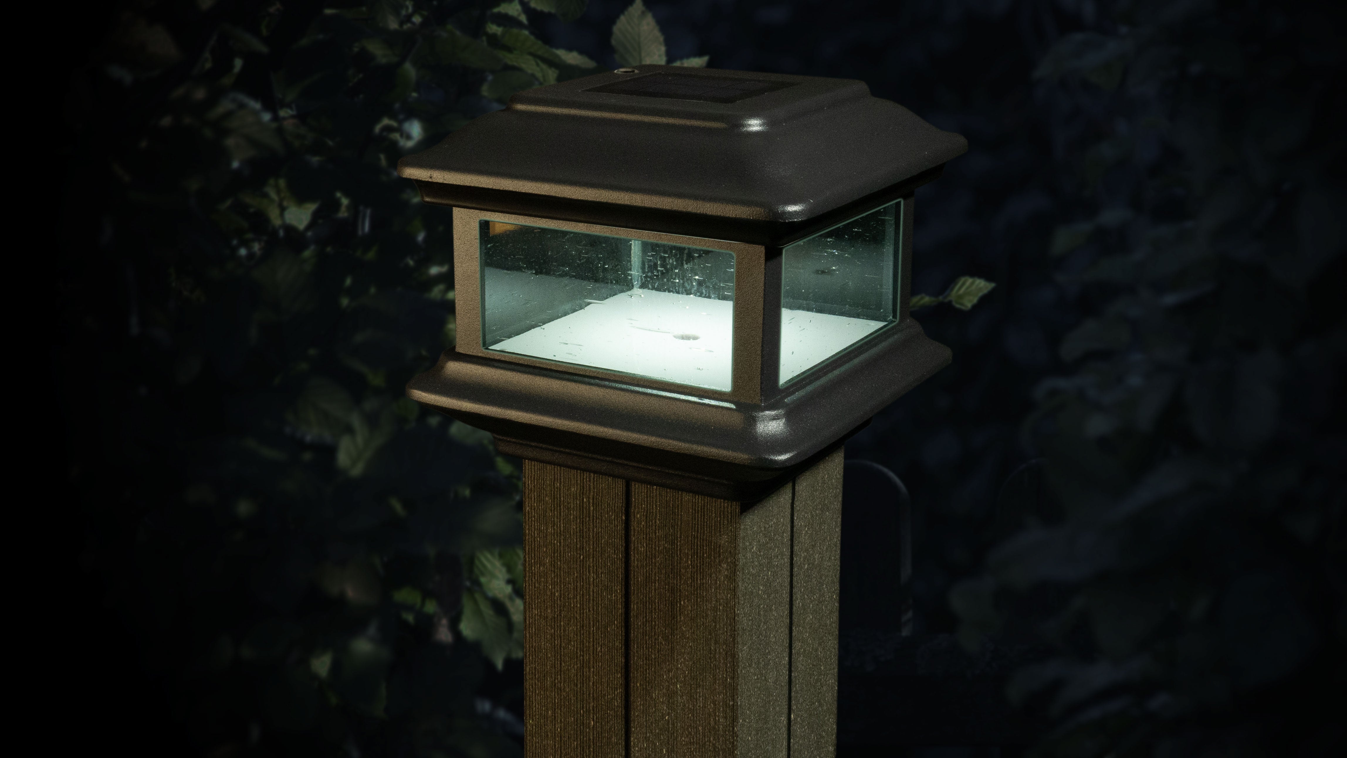 The Classy Post Cap Colonial Solar lantern, set on a wooden post amidst dark foliage, features pebbled glass panels and a square cast aluminum frame. It emits a soft white light, creating an elegant evening glow.