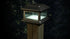 The Classy Post Cap Colonial Solar lantern, set on a wooden post amidst dark foliage, features pebbled glass panels and a square cast aluminum frame. It emits a soft white light, creating an elegant evening glow.