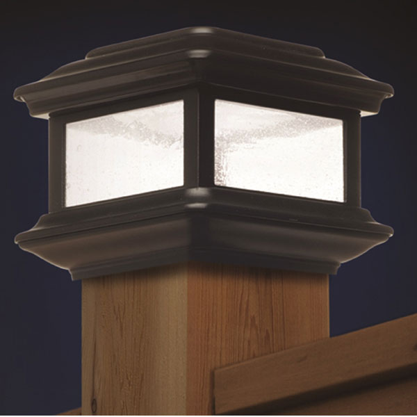 The Classy Post Cap Colonial Solar lamp is a decorative outdoor piece with square pebbled glass panels, a dark cast aluminum frame, and sits elegantly atop a wooden deck railing, glowing softly at dusk.
