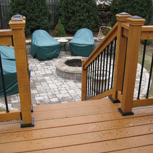 Wooden deck steps, reinforced with Post Anchor Single™, lead to a patio centered around a circular stone fire pit. Four covered outdoor chairs encircle the fire pit, framed by background evergreen trees. The deck and patio stones glisten with moisture.