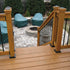Wooden deck steps, reinforced with Post Anchor Single™, lead to a patio centered around a circular stone fire pit. Four covered outdoor chairs encircle the fire pit, framed by background evergreen trees. The deck and patio stones glisten with moisture.