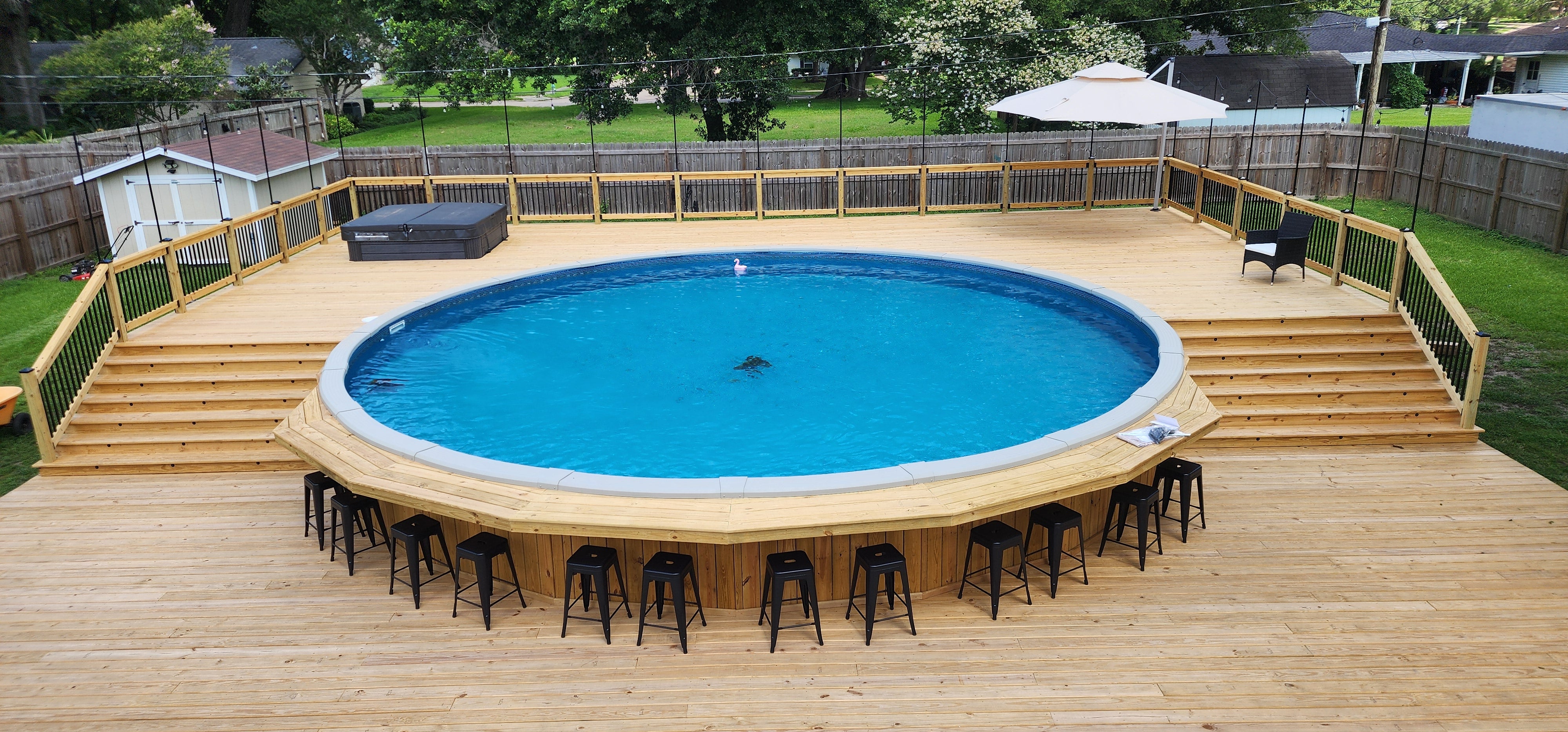 Round above-ground pool on a wooden deck with chairs around it, set in a backyard.