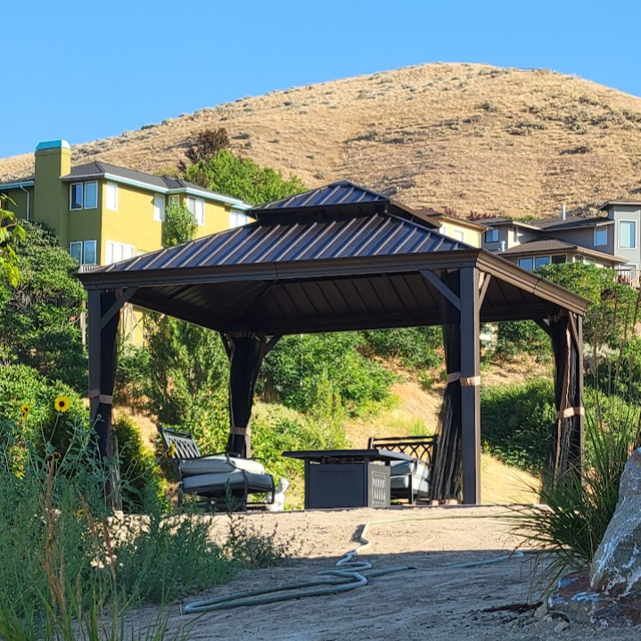 Wooden gazebo with outdoor furniture on a hillside with a building in the background