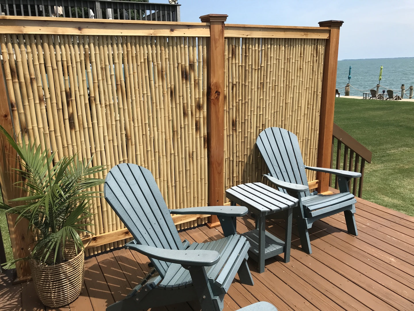 Two gray Adirondack chairs on a wooden deck with a bamboo screen and ocean view.
